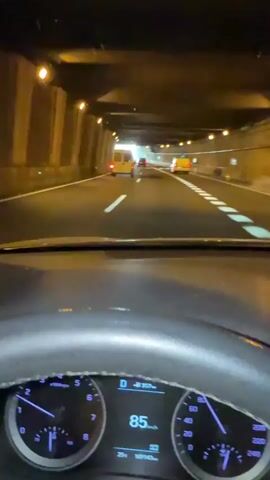 The camera is mounted on the dashboard of a car, facing forward, showing the view from inside the vehicle as it drives through a tunnel. The dashboard, with a steering wheel and part of the instrument cluster showing 86 km/h, is visible in the foreground. The tunnel walls are concrete, illuminated by lights mounted on the ceiling and sides. White lines are painted on the asphalt road, marking lanes and indicating arrows for direction. Several other vehicles, including two white vans and one yellow vehicle, are visible ahead in the distance, also driving in the tunnel. The far end of the tunnel appears bright, suggesting an exit or a section with more natural light. There's a slight glare on the windshield. The overall lighting is artificial and warm.