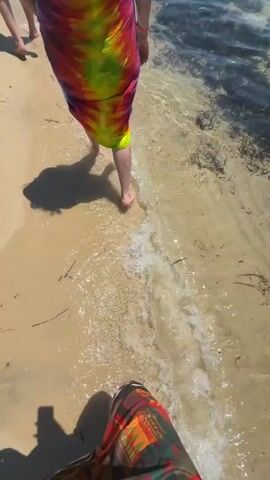 The video shows a person walking on a sandy beach towards a clear, shallow ocean. The person is wearing a colorful, flowing dress or wrap. The water is clear and shows patches of dark seaweed on the sandy bottom. In the distance, there are buildings and other people on the beach under a clear, blue sky. The camera follows the person as they walk along the shoreline.