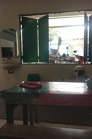 The video shows an indoor scene with a long table and several chairs. A window with green shutters is visible on the left side of the frame. A mirror is mounted on the wall above a sink. A fan is mounted on the wall below the mirror. There are several stacks of items on shelves behind the table, and a red tissue box sits on the table. A person is partially visible sitting at the table. The floor appears to be wet. The video is slightly tilted.