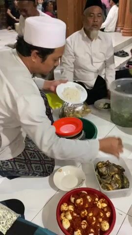 A man in a white taqiyah and white shirt is sitting on the floor, serving food from a large green bowl into a white plate. He is holding a white plate in his left hand and using a spoon in his right hand. In front of him, on the floor, are stacks of plates, a red bowl with food, and a clear plastic container with food. Another man in a white taqiyah and white shirt is sitting to the right, facing the camera, and appears to be looking at the man serving food. The environment appears to be indoors with tiled flooring and wooden walls in the background. Other people are partially visible in the background.