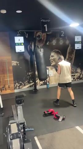 A man is punching a black punching bag in a gym. He is wearing a white tank top and black shorts, and boxing gloves. There are two screens on the wall displaying workout instructions. There is a rowing machine in the foreground. The gym has a large mural of a boxer on the wall. There are also two red boxing gloves and a timer on the floor. The man is in the middle of a boxing exercise.