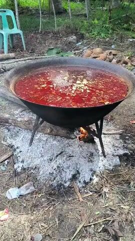 The video shows a large, black pot filled with a red, soupy substance being cooked over an open fire. The pot is supported by a metal tripod. The fire is burning underneath the pot, with visible flames and gray ash surrounding it. The environment appears to be outdoors, with trees and foliage visible in the background. There are also some discarded items, such as a plastic bottle and what appear to be dried coconut shells, scattered around the cooking area. A green plastic chair is visible in the upper left portion of the frame.