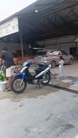 A man is washing a green motorcycle. He is standing next to the motorcycle and appears to be using a bucket and a cloth. A blue motorcycle is parked next to the green one. In the background, there is a building with a sign that says "MEBEL JEPARA INDAH". There are also some cars parked in the background. The ground is wet, indicating that the washing is in progress. A child is visible on the right side of the frame.
