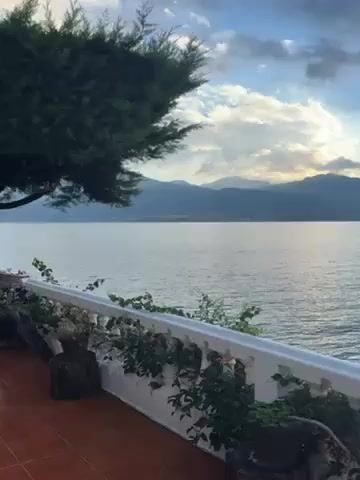 The video shows a view from a balcony overlooking a large body of water. In the distance, there are mountains silhouetted against a cloudy sky with some sunlight breaking through. The balcony has a white railing with plants growing along it. A hand reaches into the frame holding a string of lights. The lights are then hung along the railing. The water is calm with a slight ripple. The scene appears peaceful.