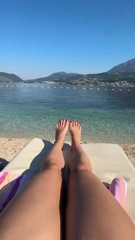 The video shows a first-person perspective of a person's legs and feet lying on a lounge chair on a beach. The person's toenails are painted red. The beach is made of small pebbles and the water is clear and blue. In the background, there is a town with buildings and mountains. The sky is clear and blue.