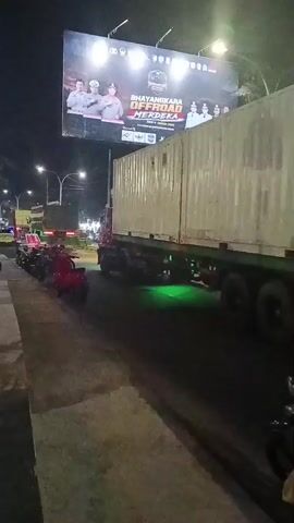 The video shows a nighttime street scene with a large billboard in the background. The billboard features images of people in uniforms and text that includes "BHAYANGKARA OFFROAD MERDEKA." In the foreground, a semi-truck with a white shipping container is driving down the street. The truck has green lights underneath the trailer and red taillights. On the side of the road, there are several parked motorcycles and a few people sitting on a low wall. Streetlights and building lights illuminate the scene. The road appears to be paved.