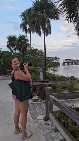 A woman is standing on a path near a beach with palm trees and a long bridge in the background. The sky is light with pink and yellow tones, suggesting it is either sunrise or sunset. The woman is holding a camera and appears to be taking a photo. She is wearing a bathing suit and a dark green bag. There is a wooden fence next to the path, and vegetation is growing along it. The water is calm, reflecting the sky.