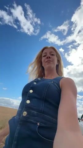 A blonde woman in a denim dress and a star necklace stands in a field with her arms outstretched. She smiles and sticks out her tongue at the camera, which is held by herself in a selfie-style shot. The background is a wide-open field of dry grass under a bright blue sky with white clouds. The sun is shining and casting light on the woman. The scene suggests a joyful and free-spirited moment.