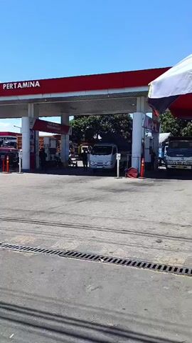 The video shows a Pertamina gas station. Several vehicles are visible at the gas station, including trucks and what appears to be a three-wheeled utility vehicle. People are also visible at the gas station, interacting with the vehicles. The gas station is brightly lit by the sun, and the sky is clear and blue. Palm trees are visible in the background. The video pans across the gas station, providing a comprehensive view of the scene.