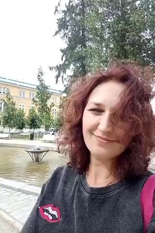 A woman with auburn, curly hair smiles at the camera. She is wearing a dark gray sweatshirt with a red and white lip patch on the front and a pink strap is visible on her right shoulder. She is standing outdoors in front of a fountain and a body of water, with trees and a building in the background. The overall setting appears to be a park or public square on a cloudy day.