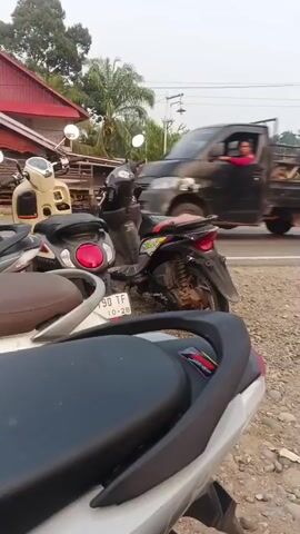 The video shows a roadside scene. In the foreground, a black motorcycle seat is visible. In the mid-ground, there are several parked motorcycles, including a silver and red one, and a black and yellow one. A grey car is parked on the side of the road, and a person in a dark headscarf and a green skirt is standing next to it. A dark colored van is driving away from the camera. Another person, wearing a blue hijab and black pants, is riding a red motorcycle and is heading towards the right side of the frame. The environment appears to be rural, with palm trees and other vegetation visible in the background. The sky is overcast.