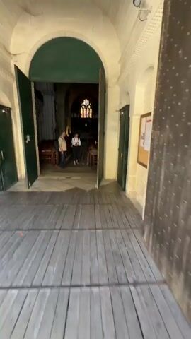 The video shows the entrance of a church. Two people are walking into the church through large, open green doors. The floor is made of wood planks in the foreground and checkered stone tiles inside the church. Rows of wooden chairs with small wooden tables are visible inside. The ceiling is arched, and stained-glass windows are visible at the far end of the church.