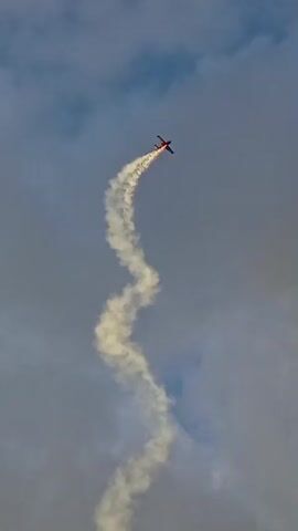 A small red airplane is performing aerobatics in a cloudy sky. The plane is leaving a white smoke trail that curves and snakes through the air. The camera is angled upwards, capturing the plane against the backdrop of gray and white clouds. The smoke trail is the most prominent feature, showcasing the complex maneuvers of the aircraft. The airplane itself is small in the frame, a tiny red speck against the vast expanse of the sky. The video appears to be a close-up shot, focusing on the action of the airplane and its smoke trail.
