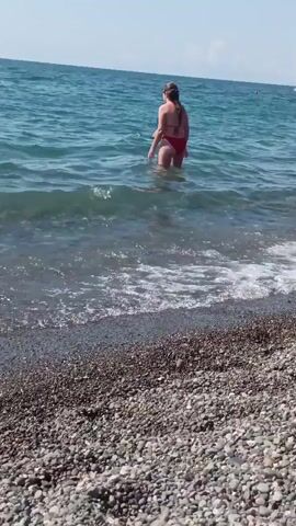 A woman in a red bikini is standing in the ocean near a pebbled beach. She is bending forward, reaching into the water with her hands. Her hair is pulled back, and she is wearing sunglasses. The water is blue and appears calm with small waves. In the background, another person is partially visible behind her in the water.