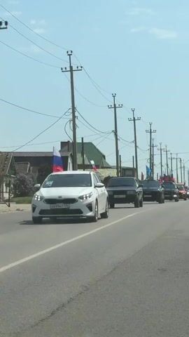 The video shows a procession of cars driving down a paved road on a sunny day. The cars are of various makes and models, and most have Russian flags attached to them. The road is lined with utility poles and there are buildings in the background. The camera appears to be in a moving vehicle.