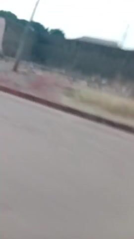 The video shows a blurry view from a moving vehicle driving on a paved road. On the right side of the road, there are houses and other structures behind a curb. The ground is reddish dirt. In the distance, a tower structure is visible against the sky. The sky is a pale color, possibly indicating early morning or late evening. The video is shaky and the focus is not sharp.
