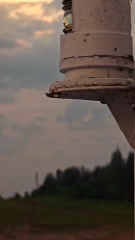 This is a close-up, low-angle shot of a white, weathered buoy light against a dramatic sky at dusk. The light fixture, with its round glass lens, is the primary subject, showing signs of age with peeling paint and rust. Several fine strands, possibly spider webs, are visible hanging from the underside of the light's platform, catching the light.

The background is softly blurred, creating a sense of depth. It depicts a rural landscape with a gentle slope of green grass leading to a line of dark trees against a gradient of twilight sky. The sky itself is a mix of muted grays, blues, and hints of warm yellow or orange from the setting sun, suggesting a peaceful, transitional time of day. The overall mood is one of quiet solitude and perhaps a touch of melancholy due to the weathered appearance of the buoy light and the approaching darkness.

There is no visible text in the media file. The scene appears to be set in a natural, possibly coastal or lakeside environment in Vorogovo, Russia, given the context provided. The activity is static; the buoy light is stationary, and the image captures a moment in time.