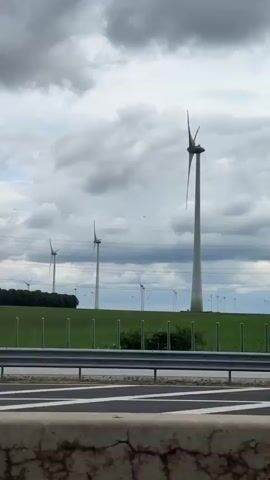 From a moving vehicle on a highway, a view is captured of a field of wind turbines under a cloudy sky. The turbines are white and stand tall in the green field, with power lines visible in the background. A metal guardrail runs alongside the highway. The camera briefly passes some greenery.