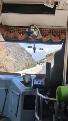 The video provides a first-person perspective from inside a bus, looking out the front windshield at a scenic mountain road. The dashboard of the bus is visible in the foreground, with a navigation screen and various controls. Outside the windshield, a winding road curves through a mountainous landscape. On the side of the road, a white SUV is parked, and several people are standing near it. Further down the road, another vehicle appears to be driving. The background is dominated by majestic snow-capped mountains under a clear blue sky. The bus is moving, as indicated by the slight motion blur and the changing perspective of the road. The interior of the bus has decorative trim along the top of the windshield and a green sleeping mat is visible on a seat.