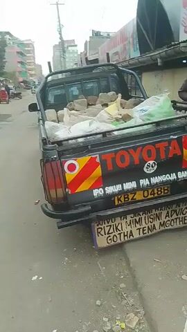 The video shows the back of a black Toyota pickup truck parked on a street. The truck bed is filled with bags and large tan bricks. The truck has red and yellow diagonal stripes, the word "Toyota" in red, and a license plate that reads "KBZ 048B". Below the license plate, there's a small sign with text in Swahili. The street appears to be in a built-up urban area, with buildings on both sides, including one with a sign that reads "BUTCHER". There are also power lines overhead and some other vehicles visible down the street. A hand briefly appears on the right side of the frame.