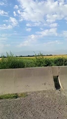 The video shows a concrete barrier with an opening in the middle, which appears to be a culvert or drainage channel. Inside the opening, there is trash, including a blue plastic bottle. Beyond the barrier, there is vegetation and a field, with mountains in the distance under a cloudy sky. The camera is moving from left to right, capturing the scene.