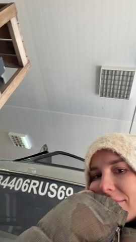 A young woman is featured in a close-up, selfie-style video shot indoors. She wears a cream-colored, fuzzy winter hat and a grey, quilted jacket, suggesting cool temperatures. Her expression is calm and she offers a slight smile to the camera. Behind her, a grey boat or jet ski is partially visible, bearing the registration number "TB4406RUS" on its side. The interior setting appears to be a garage, workshop, or storage facility, characterized by a white paneled ceiling and fluorescent strip lighting. A wooden shelf or structure is visible in the upper left corner of the frame. The woman shifts her head slightly during the short clip.