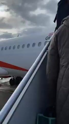 The video begins with a close-up shot of people boarding an airplane. One person has a gray backpack. The view then shifts to a scene of an EasyJet airplane on the tarmac, seen from the wing of another plane. The sky is cloudy. The EasyJet plane is in motion, seemingly taxiing. The ground is wet, possibly from rain.