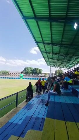 The video shows a soccer field from the stands. The stands are covered with a green awning. The seats are painted blue and yellow. People are sitting in the stands, watching the soccer players practice on the field. The field is green and surrounded by a blue and white wall. There are trees in the background and a blue sky with white clouds overhead. A green, inflatable coconut balloon is attached to the field’s wall.