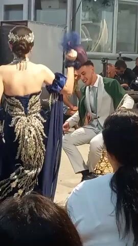 The video shows an outdoor scene, possibly a celebration, with several people present. In the foreground, partially obscured by spectators, is a man in a light-colored suit with a green sash, seated and appearing to be interacting with someone off-camera. To his left, a woman in an elaborate dark blue and gold dress stands facing away from the camera. Behind them, a man in a yellow t-shirt sits on a chair, looking towards the seated man. Further back, other individuals are visible, some seemingly observing the scene. A large black speaker is positioned to the left of the frame. The ground appears to be dirt or gravel. The overall atmosphere suggests a lively event.