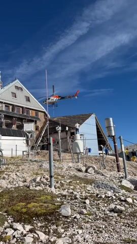 A helicopter is shown in flight near a mountain facility. The facility appears to be an alpine research station or lodge, constructed with wood and metal. There are several meteorological instruments visible, including what look like rain gauges and weather sensors. A few people are present on the roof of a structure to the right of the main building. The environment is rocky and barren, with sparse vegetation. The sky is blue with some wispy clouds.