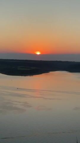 The video captures a serene sunset over a calm body of water. The sun, a bright orange orb, is partially obscured by a thin line of clouds on the horizon, casting a warm glow across the sky and reflecting on the water's surface. A dark, silhouetted landscape borders the water, with hints of rolling hills or vegetation. A small boat is visible in the distance, leaving a faint wake on the tranquil water. The overall atmosphere is peaceful and picturesque, emphasizing the natural beauty of the sunset.