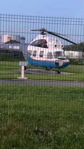 This video shows an outdoor scene at what appears to be an airfield or aviation museum. A fence with green posts and mesh runs across the foreground, partially obscuring the view. Behind the fence, several aircraft are visible. On the left, a white and blue helicopter with the registration RA-15619 is parked on a pad. To the right of the helicopter, a biplane with a visible propeller and landing gear is parked. Further to the right, a smaller twin-engine propeller plane with a white fuselage and blue stripe is also parked. The background includes grassy areas, buildings, and what appear to be aircraft hangars or storage facilities. The sky is visible in the upper portion of the frame and suggests it may be dusk or early morning, with soft, diffused lighting. There are no people clearly visible in the frame. The overall atmosphere is static, with the aircraft appearing to be stationary.