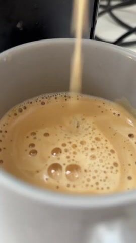 A close-up shot shows coffee being poured into a white mug. The coffee is light brown with a foamy top layer filled with small bubbles. The stream of coffee is a darker brown and is creating ripples on the surface of the existing liquid. A black coffee machine is partially visible in the upper left corner of the frame. The background is out of focus, but appears to be a white surface with a black cord.