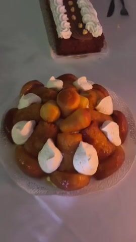 The video shows a close-up view of several desserts on a table. In the foreground, a large, circular cake or pastry is filled with small, golden-brown, rum-soaked cake balls and topped with dollops of whipped cream. In the background, a rectangular chocolate cake is decorated with white frosting piped in a wave pattern, and small nuts or candies. A third dessert, a round cake with layered frosting and sprinkled nuts, is also visible. Cutlery, including a fork and knife, are present on the table, suggesting a meal or celebration. The lighting is dim, casting a warm glow on the desserts.