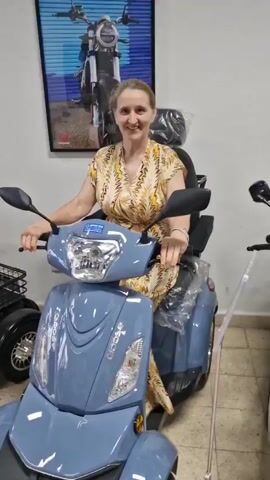 A woman is sitting on a blue motorized scooter in what appears to be a showroom. She is wearing a patterned dress and smiling at the camera. There are other scooters visible in the background, as well as a framed picture of a scooter on the wall. The scooter has three wheels, a large seat, and mirrors. There is plastic wrap on some parts of the scooter.