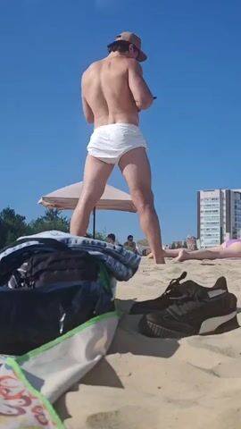A man wearing a baseball cap and white shorts stands on a sandy beach, facing away from the camera. He is holding a phone in his right hand. In the foreground, a bag and a pair of black sneakers are placed on the sand. The sky is clear and blue. In the background, a beach umbrella, some trees, a building, and other beachgoers are visible. The video is a still shot.