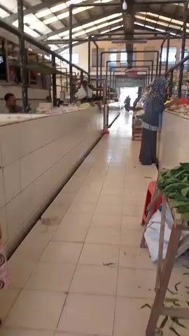 The video shows a person walking down an aisle in an indoor market. The aisle is lined with stalls on either side, some of which appear to be empty. The floor is tiled, and the ceiling is made of metal with translucent panels allowing light to enter. A woman wearing a headscarf and a long skirt is visible at a stall on the right side of the aisle. She appears to be interacting with a vendor, or looking at products. Other people are visible in the distance at the end of the aisle. The stalls contain various items, including what appear to be produce and packaged goods. The overall impression is of a relatively quiet market, possibly early in the day or during a less busy period.