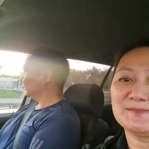 Two people are in a car. The person on the left is a man wearing a blue shirt and a seatbelt. He is looking out the window. The person on the right is a woman with dark hair wearing a black hoodie. She is looking at the camera and speaking. The car appears to be moving. The environment outside the car is visible through the windows and appears to be a road with some buildings and trees. The lighting suggests it is daytime, possibly late afternoon.
