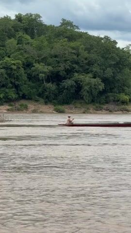 A long, narrow, maroon boat with a person wearing a light-colored hat and clothes is moving across a wide, muddy river. The person is seated near the front of the boat, holding onto a tiller that controls a small green outboard motor at the back. The boat is leaving a slight wake behind it.

The river is flanked by a steep, densely forested bank. The trees are a vibrant green, suggesting a lush environment. The terrain beyond the immediate riverbank rises into rolling hills covered with more trees. The sky is overcast with visible clouds, indicating a cloudy or potentially rainy day. The overall impression is a serene, natural landscape, likely in a rural or undeveloped area.

The image appears to be taken from another boat or from the riverbank, capturing the scene from a side-on perspective. The lighting suggests it could be late morning or afternoon, given the diffused light. There is no visible text in the media. The setting strongly suggests a location with a large river flowing through a heavily forested region, consistent with the provided location context of Don-Kao, Laos.