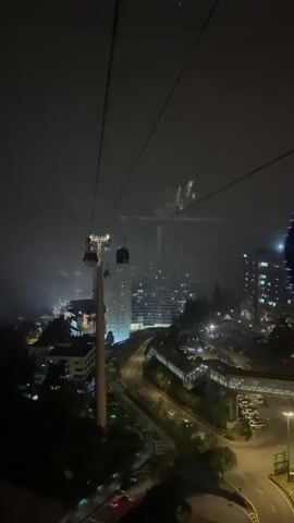The video shows a night view from a cable car looking down onto a lit-up city. Several cable cars are visible on their wires. A tall tower and other buildings are prominent in the cityscape. A winding road with cars is also visible, along with a parking lot. The sky is dark and appears to be foggy or misty. The video is shot from inside a cable car, with the wires running upwards.