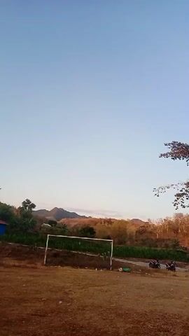 The video shows an outdoor scene with a clear sky and rolling hills in the background. In the foreground, there is a dirt field with a soccer goal. A few motorcycles are parked on a road to the right of the goal. The overall impression is of a rural landscape in the daytime.