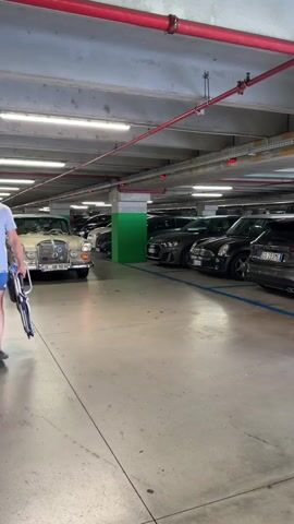 The video shows a man in a parking garage, carrying a folded chair and a bag. He is wearing a light-colored t-shirt, blue shorts, and sandals. He appears to be walking towards a vintage beige Mercedes-Benz sedan, which is parked in the foreground. The car has chrome accents and a license plate that reads "UL UR 90 H". Other cars are visible in the background, including a dark gray SUV and a black Mini Cooper. The parking garage has concrete ceilings with red pipes and fluorescent lighting. The scene suggests a warm, sunny day, typical of summer in Italy, given the casual attire of the man and the location of Limone sul Garda. The man's expression is neutral, and the overall atmosphere is mundane, focused on the act of moving between vehicles.
