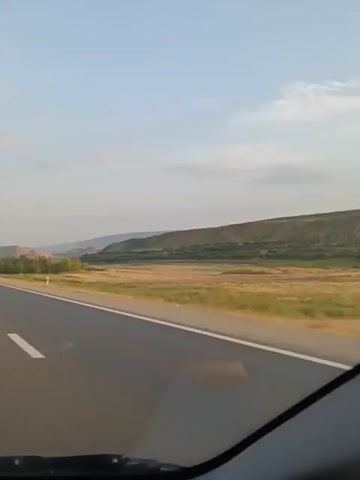 The video shows a view from a moving vehicle on a highway. The road is paved and has white lane markings. On the right side of the road, there is a grassy field with some trees and hills in the background. The sky is clear and blue with a few scattered clouds. There are other vehicles visible on the road ahead.