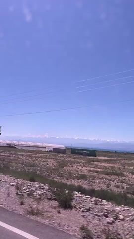 The video captures a roadside view under a clear blue sky, with a bright sun. The foreground shows a rocky, gravelly area next to a paved road, with sparse, dry vegetation. In the middle ground, there are several large, white greenhouse structures lined up. Beyond the greenhouses, a vast, shimmering body of water is visible, likely a lake, with a range of snow-capped mountains in the distance. Overhead, power lines stretch across the frame, supported by utility poles. The scene appears to be a landscape shot from a moving vehicle, as indicated by the slight blur and the perspective shift.