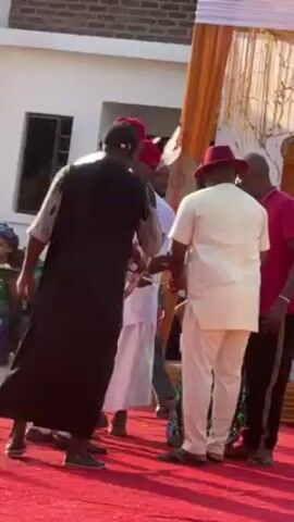The video captures a lively outdoor celebration, likely a wedding, taking place on a red carpet under a decorated canopy. 

Several men dressed in traditional African attire engage with a microphone, while people dressed in formal and casual clothing mingle around the tent.  

A woman in a bright pink outfit with a designer tote bag walks past the camera. White tents are partially visible in the background.