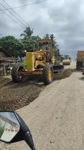 A road construction scene is depicted, with heavy machinery at work. In the foreground, the side mirror of a motorcycle offers a partial view of its handlebars. A yellow grader, a large piece of construction equipment with a blade, is positioned on the left side of the frame, actively working on the dirt road, its tires leaving tracks in the loose material. Further down the road, a yellow steamroller is compacting the surface, followed by a dump truck. A person on a motorcycle is riding past the dump truck. The environment appears to be a rural or semi-urban area with scattered buildings and palm trees visible in the background under a cloudy sky. The road itself is unpaved and appears to be undergoing repairs or resurfacing.