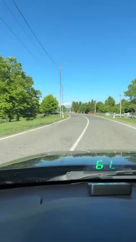 The video shows a first-person view from inside a car traveling on a road. The road is paved and has a white dividing line. There are trees and greenery on either side of the road. Overhead power lines are visible. The sky is blue and clear. A digital display on the car's dashboard shows the number "6" which then changes to "0".