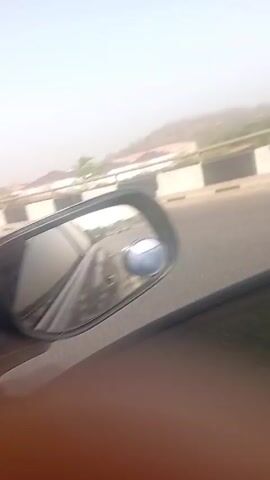 From a vehicle, the camera captures the view from the passenger window as the car moves along a highway, revealing the passing scenery. Visible is a section of the road separated by a concrete barrier with alternating black and white stripes.  In the background, residential buildings, partly obscured by a hazy atmosphere, sit on an elevated surface. An unfinished multi-story building, still surrounded by scaffolding, stands out in the distance. The sky is clear and bright, suggesting daytime, and the car’s side mirror is visible, reflecting the interior of the vehicle and a small portion of the outdoor scenery. The text 'OBJECTS IN MIRROR ARE CLOSER THAN THEY APPEAR' is seen in the side mirror.