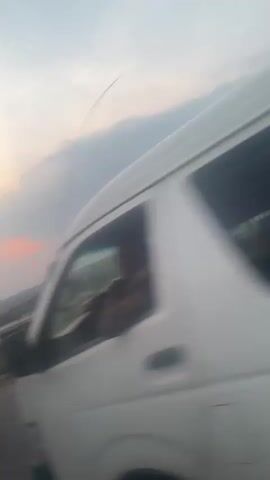 The video shows a landscape view from a moving vehicle. The foreground contains the road and a strip of green vegetation. In the background, there's a view of a town or village, with buildings and trees, all under a cloudy, early evening sky with hints of orange from the setting sun. A large tanker truck passes close to the camera, temporarily obstructing the view. The camera angle is slightly tilted.