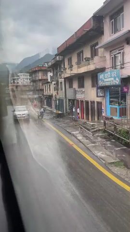 The video captures a street view from a moving vehicle on a wet, overcast day in Chandragiri, Nepal. The asphalt road is slick with rain, reflecting the grey sky, and features a distinct yellow line.

Buildings, mostly multi-story and appearing somewhat aged, line both sides of the street. Many ground floors serve as shops, some with closed or partially open shutters. Visible text on signs includes "नयाँ घडी घर" (Naya Ghadi Ghar - New Watch House) on a blue sign and "अग्नि सरज क्लास" (Agni Saraj Class) with "हेयर कट" (Hair Cut) on a green shop sign, indicating a barbershop or salon.

In the initial frames, a silver Suzuki Swift car (license plate बा ८ च ८२५८) drives past in the opposite direction, creating a spray of water. A dark blue/purple motorbike is parked on the side of the road facing the camera. Further down, another red/pink motorbike is briefly visible.

A few people are seen on the sidewalks. At one point, a man in a dark jacket stands with two children near the barbershop, looking towards the street. Mountains, partially obscured by mist or clouds, form the backdrop in the distance. Power lines crisscross above the street.